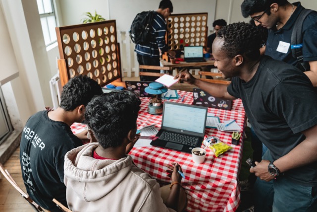 Hack Day participants working on laptops around a table while a host leans in to help